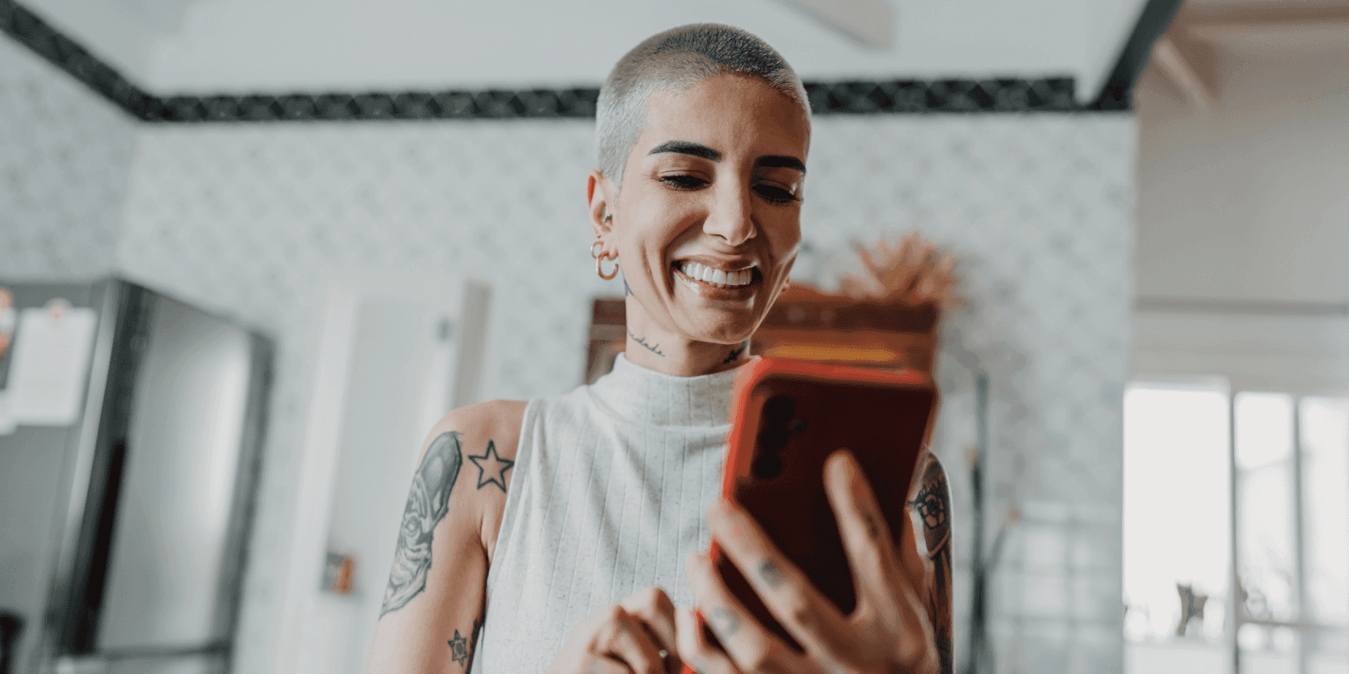 A woman with a shaved head and tattoos scrolling on her red phone. She is smiling lightly.