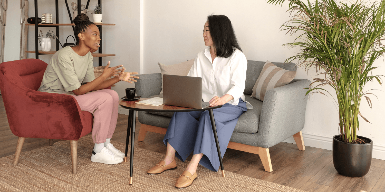 A therapist and client talking. The therapist has a laptop for notes, and the client is gesturing with her hands. Both are smiling slightly.