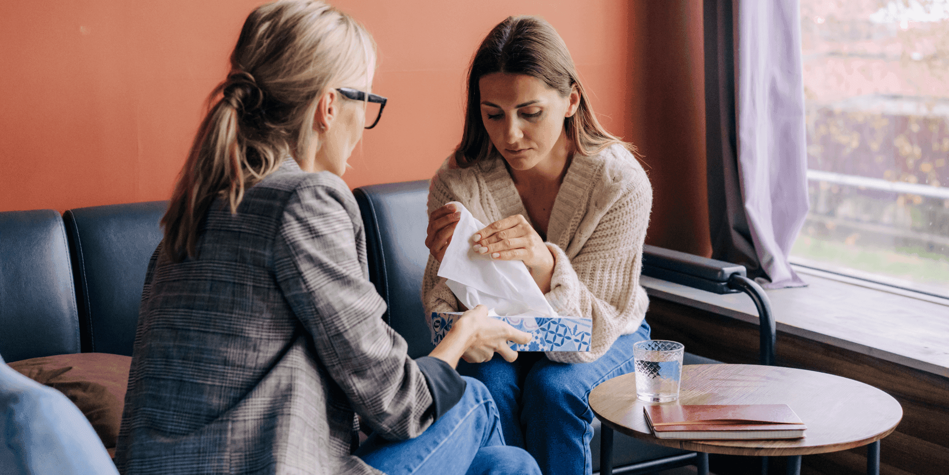A therapist hands a box of tissues to a young woman who looks upset.