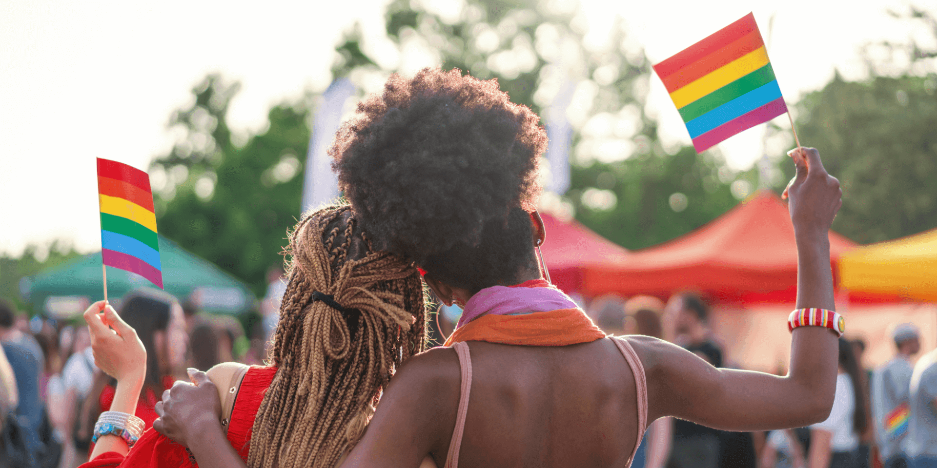 Two women standing side by side with their arms around each other, each holding an LGBTQ+ Pride flag.